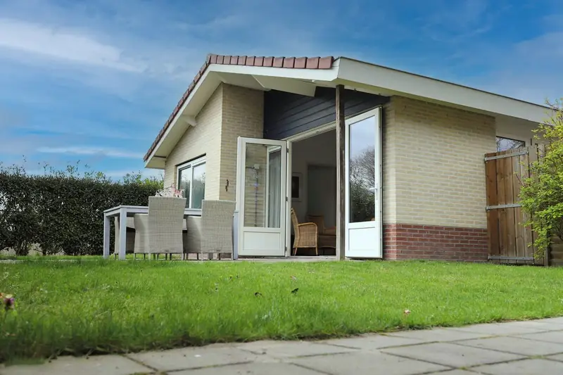 De achterkant van huisje 2, met open terrasdeuren, de tuintafel met stoelen en het gazon.