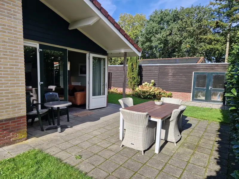 Het terras van huisje&nbsp;2: een houten tafel met vier rieten stoelen, bij de open terrasdeuren een bistrotafeltje, en achter het gras de schuur.